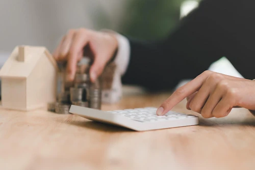 Main manipulant une calculatrice prÚs de piÚces empilées et d'une maquette de maison en bois.