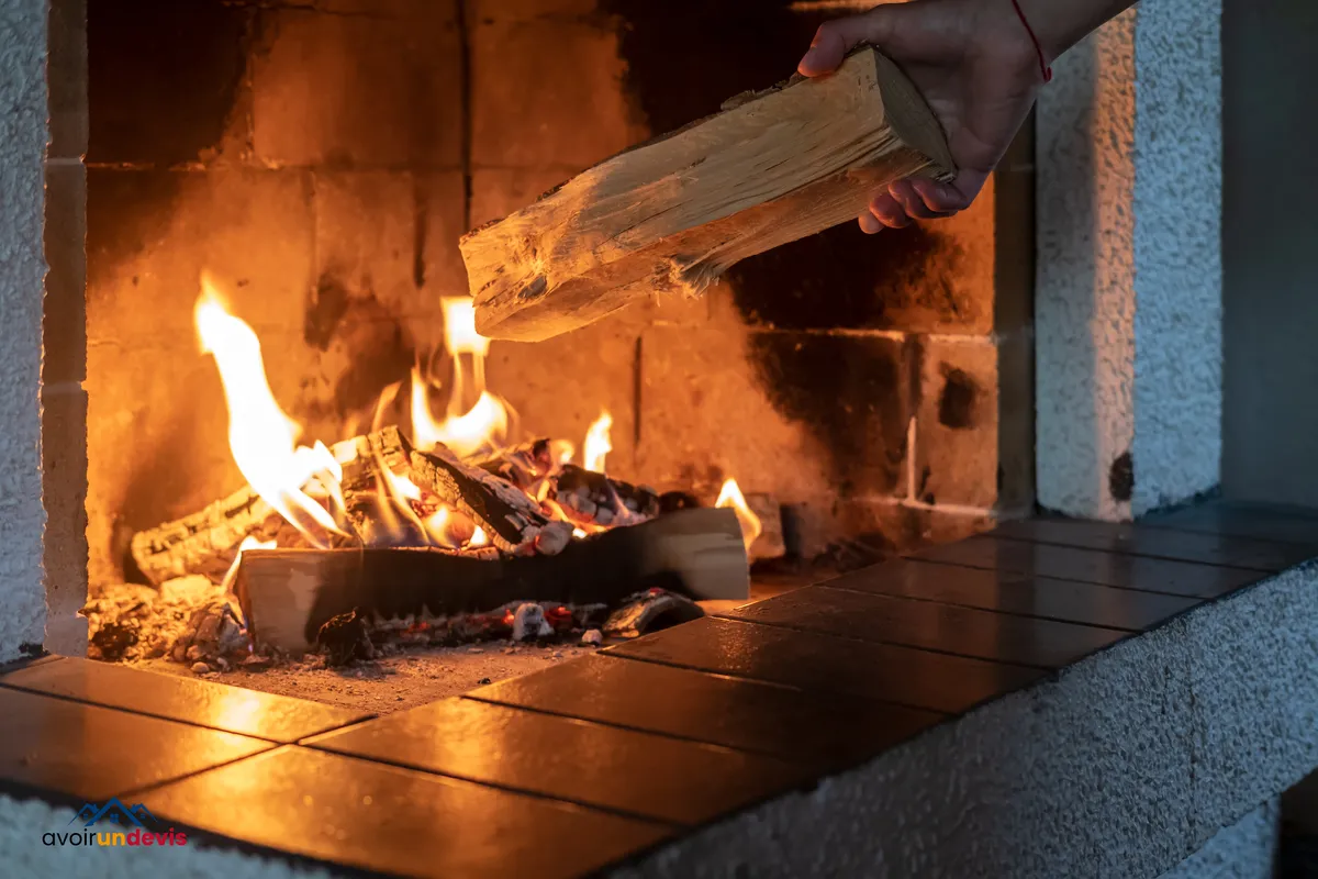 Main plaçant une bûche dans une cheminée allumée avec des flammes visibles et des braises incandescentes, à l'intérieur d'une maison.