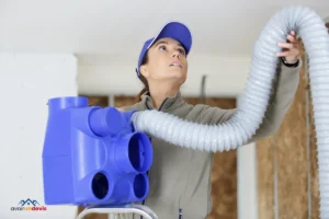 Une professionnelle en vêtements de travail et casquette examine attentivement l'installation d'un système de ventilation. Elle tient un conduit flexible, le positionnant probablement dans le cadre de l'installation ou de la maintenance du système. Son regard est concentré vers le haut, indiquant qu'elle est en train d'évaluer le travail à effectuer ou de vérifier l'installation.