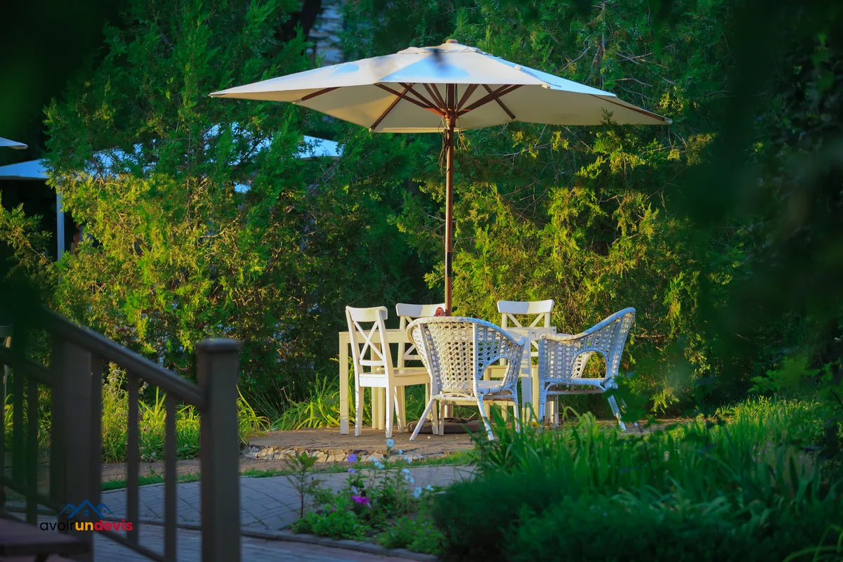 Un coin paisible de jardin est aménagé avec une grande table entourée de chaises blanches sous un parasol ouvert, offrant un espace ombragé et accueillant. L'environnement est verdoyant, avec des arbres et des plantes luxuriantes qui entourent la terrasse, créant une atmosphère tranquille et isolée idéale pour la détente ou les repas en plein air.